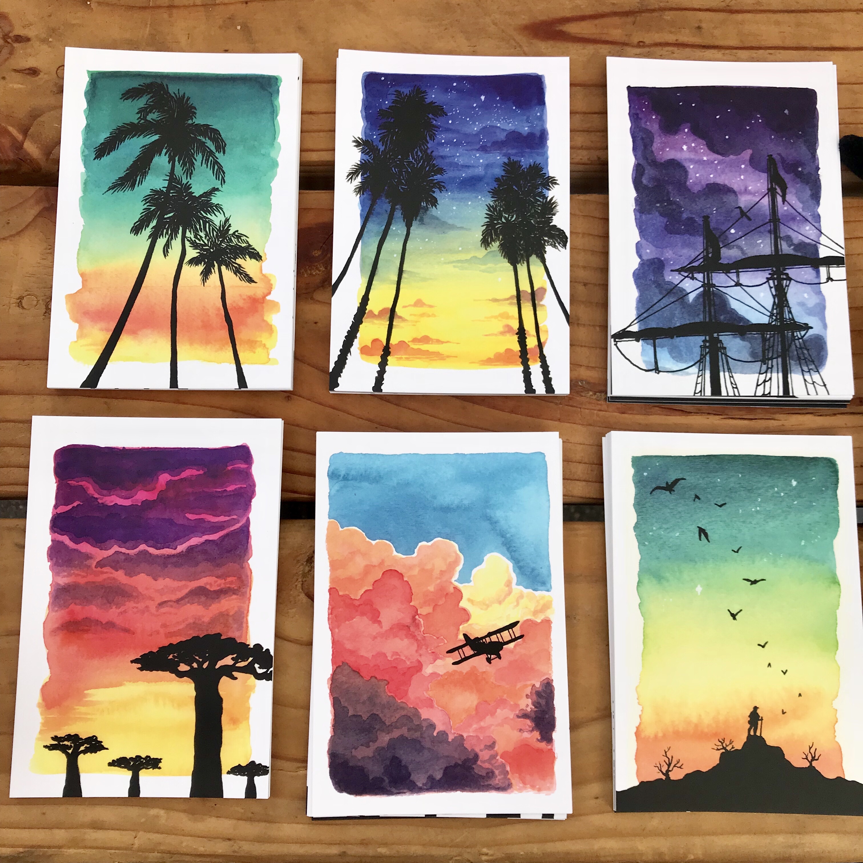 A selection of vibrant postcards featuring watercolor paintings of sunrises and sunsets with silhouettes of trees, tall ships, mountains, and other organic shapes in the foreground.