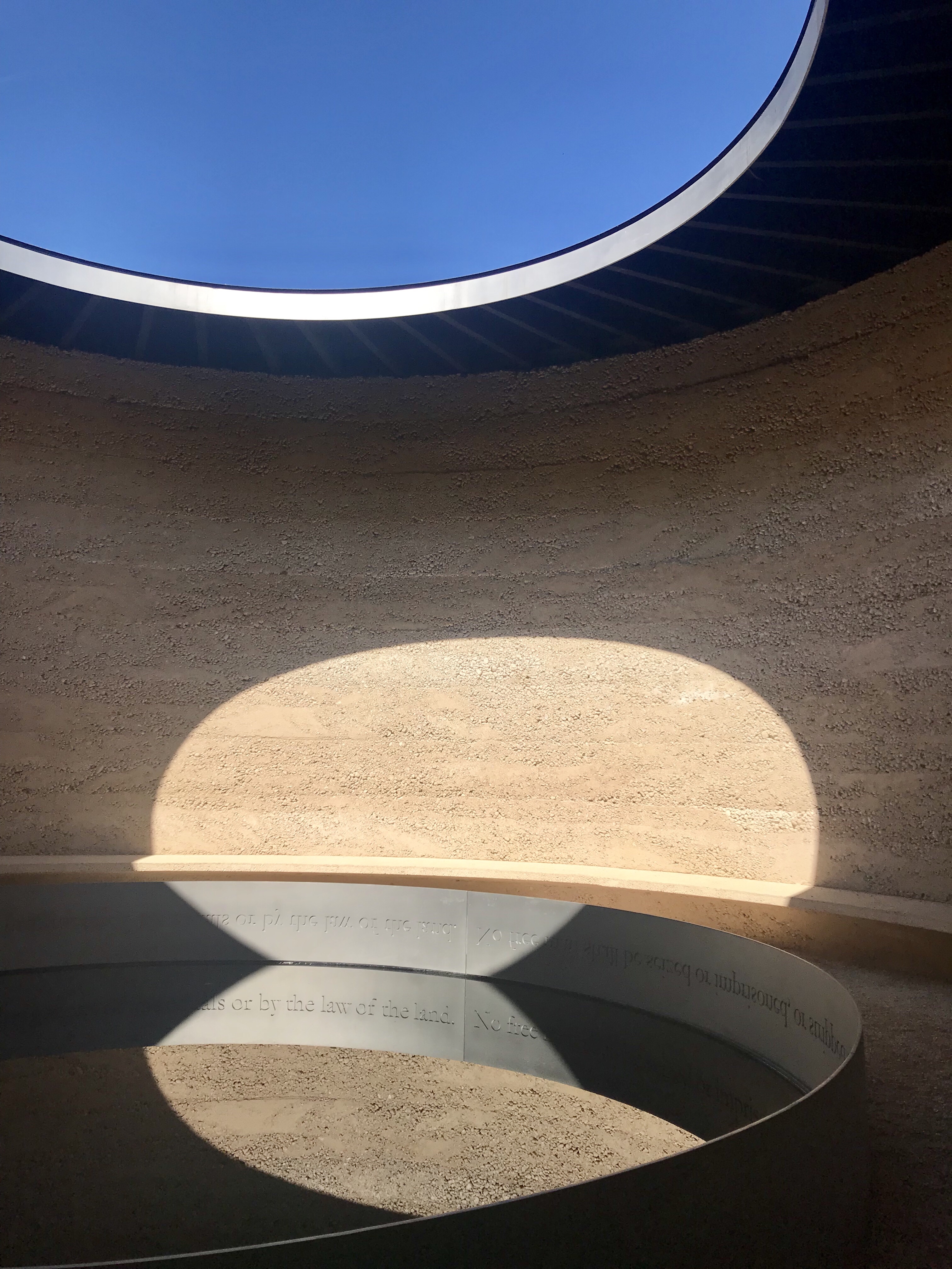 A circular space with a pond in the center and an open circle in the roof. Text written upside down around the rim of the pool is reflected right-side up in the water. The walls are pale and rough, the sky is blue.