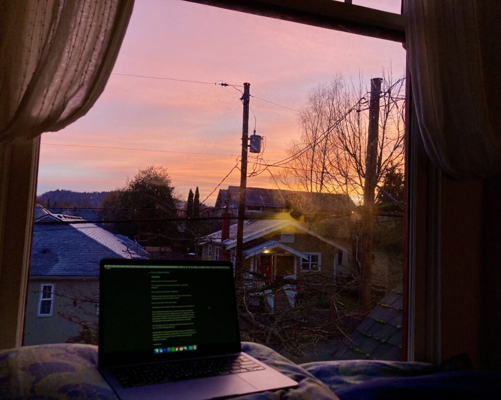 A laptop resting on a pillow in front of a window. There's a luminous pink sunrise glowing on the horizon.