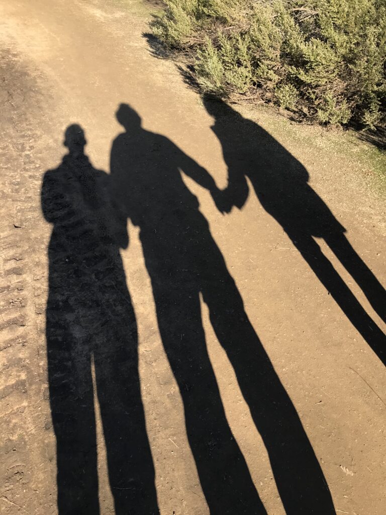 Three elongated human shadows, holding hands and walking along a dusty trail.