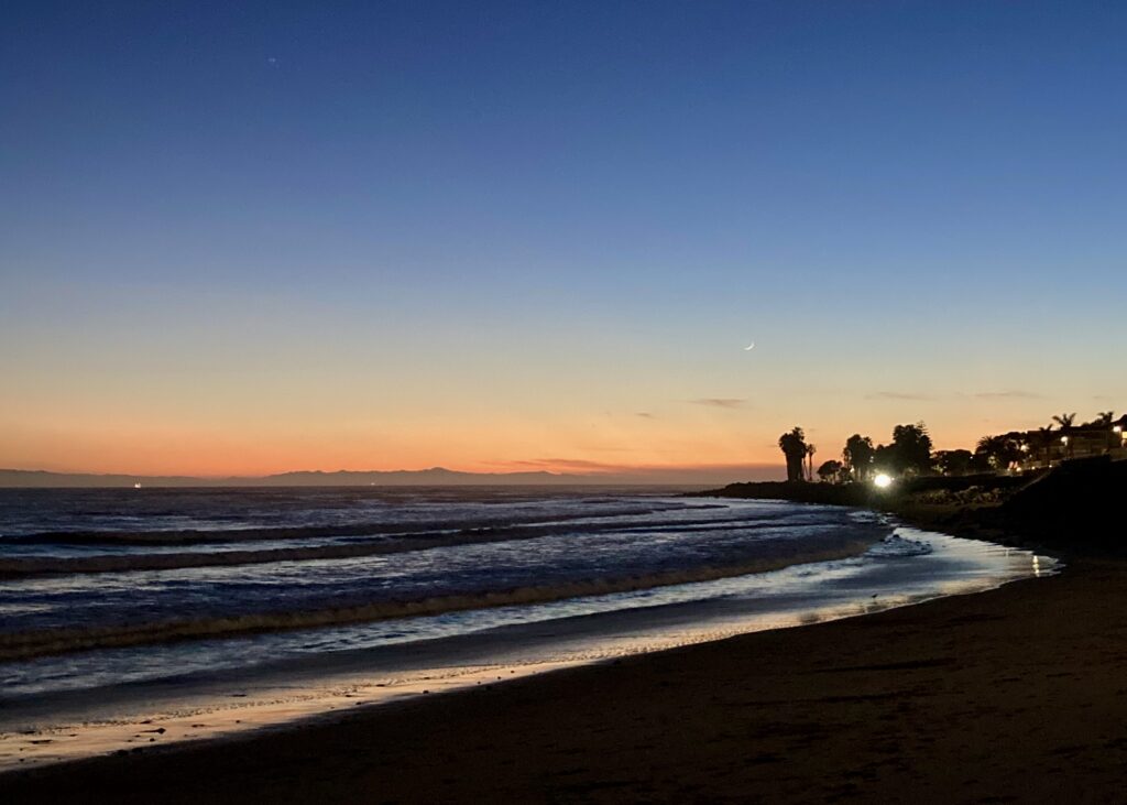 A glowing beach at sunset. There are shallow, reflective waves moving up the shore. The sky is apricot at the horizon grading slowly into deep blue. On the right, a headland with palm trees silhouetted against the sky. There's a tiny sliver of moon rising.