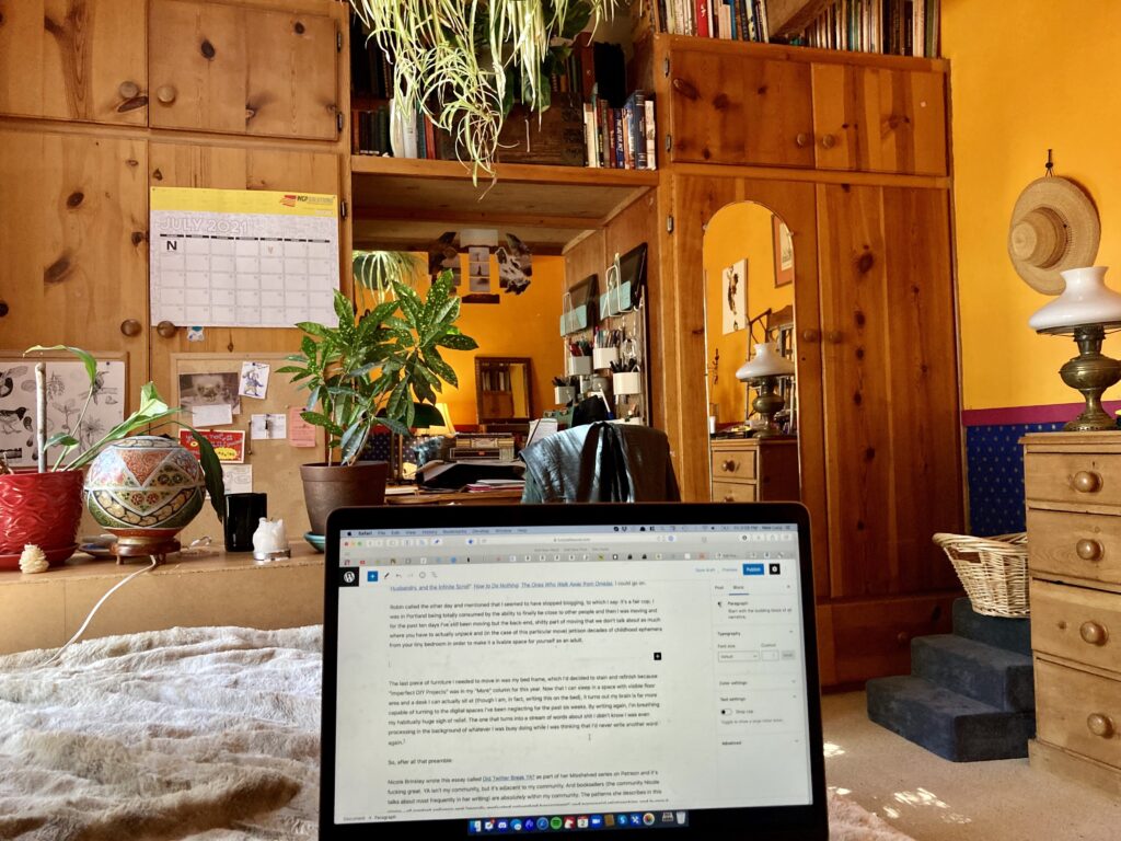 A POV photo from Lucy's bed. Her laptop is open in the forground with this blog post on it. There are wooden cabinets and a lot of books and orange walls and houseplants. It's cozy and warm.