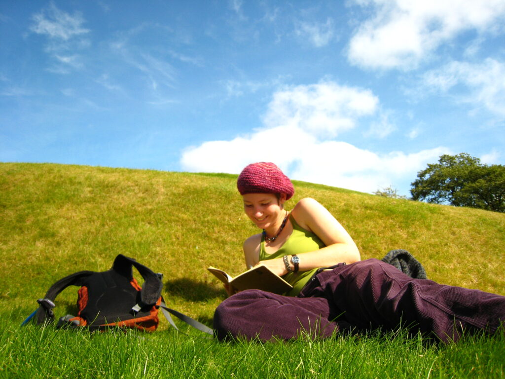 A saturated photo of Lucy on a green hillside on a sunny day. She's lying on the grass reading a book, a small orange backpack on the turf next to her.