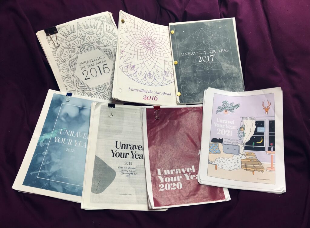 A collection of printed workbooks with creative covers on a purple bedsheet, each labeled with "Unravel Your Year" and then a date stretching from 2015 to 2021.