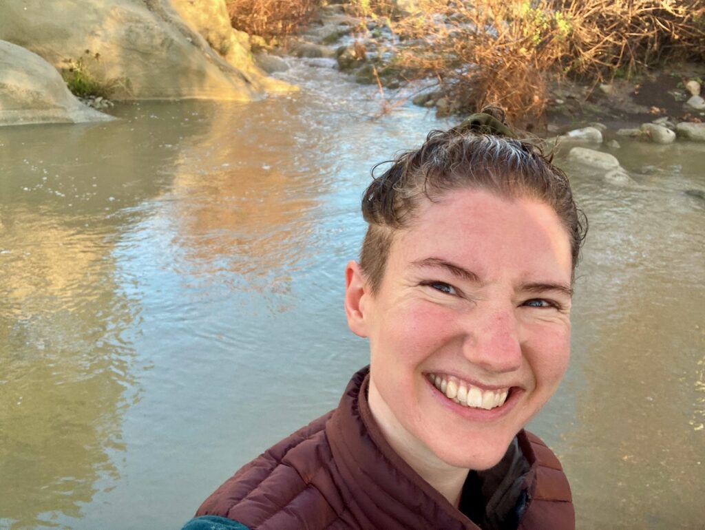 Lucy, a young white woman with an sodden undercut, grins at the camera in front of a muddy river swimming pool.