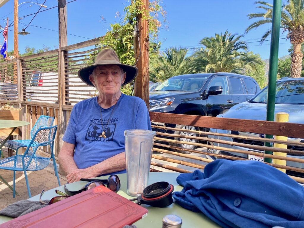 A photograph of Lucy's dad, Peter, wearing a blue tshirt and a sun hat and smiling at the camera. He's sitting on a patio in the shade.