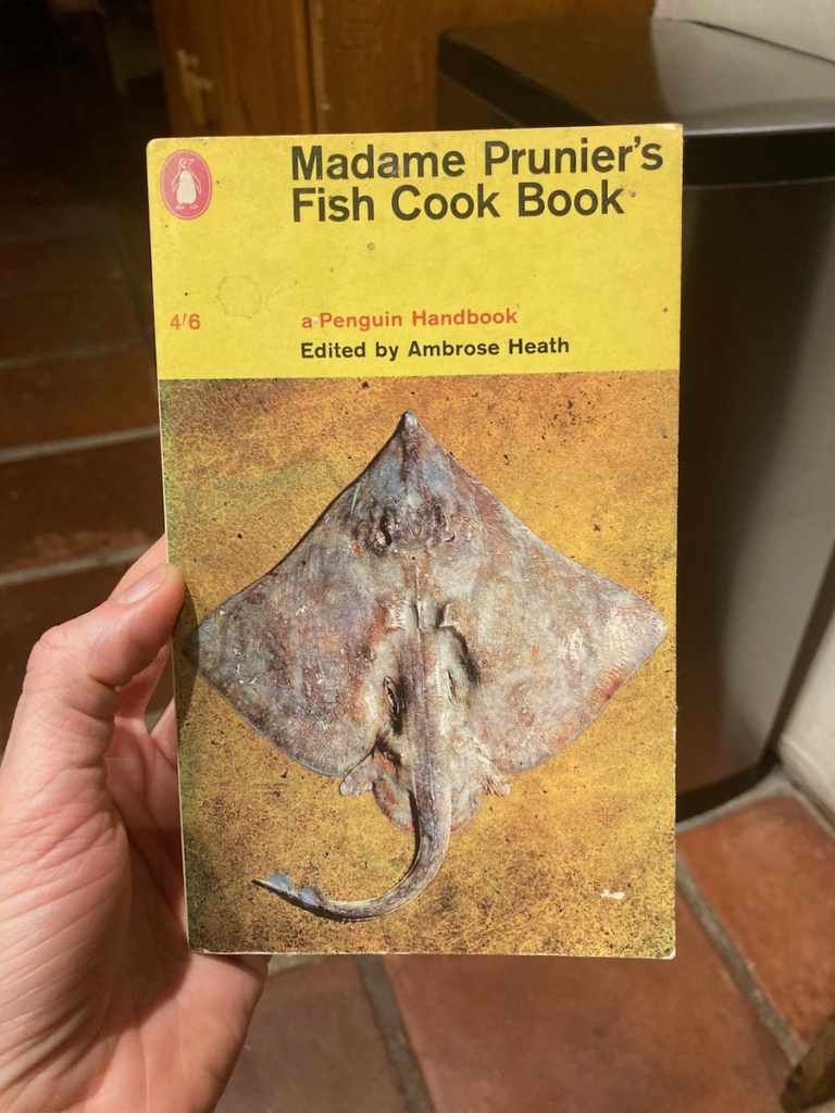 A photo of a hand holding a paperback book titled Madame Prunier's Fish Cook Book. There's sickly yellow background color to the text and a large, unappetizing photograph of a sting ray below it.