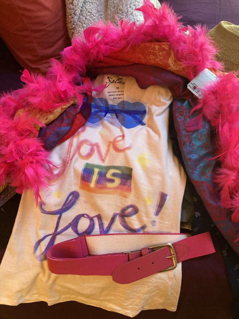 A collection of bright pink belts, a feather boa, a pair of blue heart-shaped sunglasses, and a white shirt that says love is love.