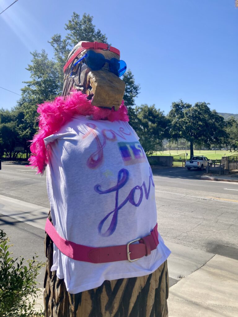 A carved wooden statue of a bald eagle wearing heart-shaped glasses, pink feather boa, and love is love t-shirt.