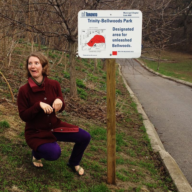 Lucy panting like a dog beside a sign that reads “Trinity-Bellwoods Park. Designated area for unleashed Bellwoods.”