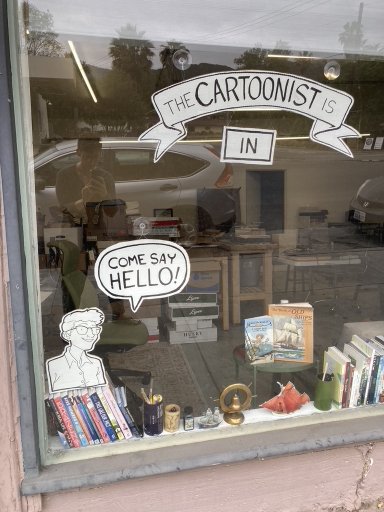 A photo of Lucy's actual studio window with a real sign saying "The cartoonist is in." There's also a drawing of her saying "Come say hello!"