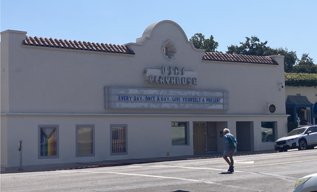 The Ojai Playhouse, a Spanish-style building whose marquee says "Every day, once a day, give yourself a present." There's a pride flag in one of the poster windows.