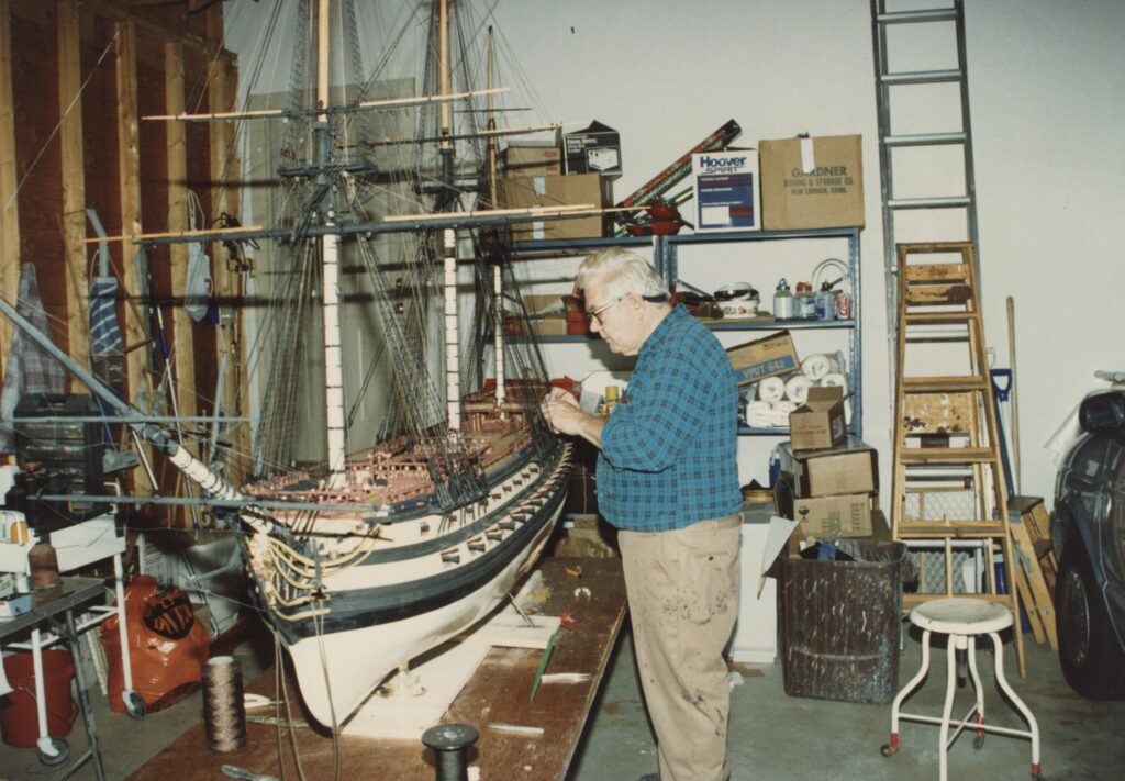 William Hitchcock at work beside a massive model ship on a workbench.